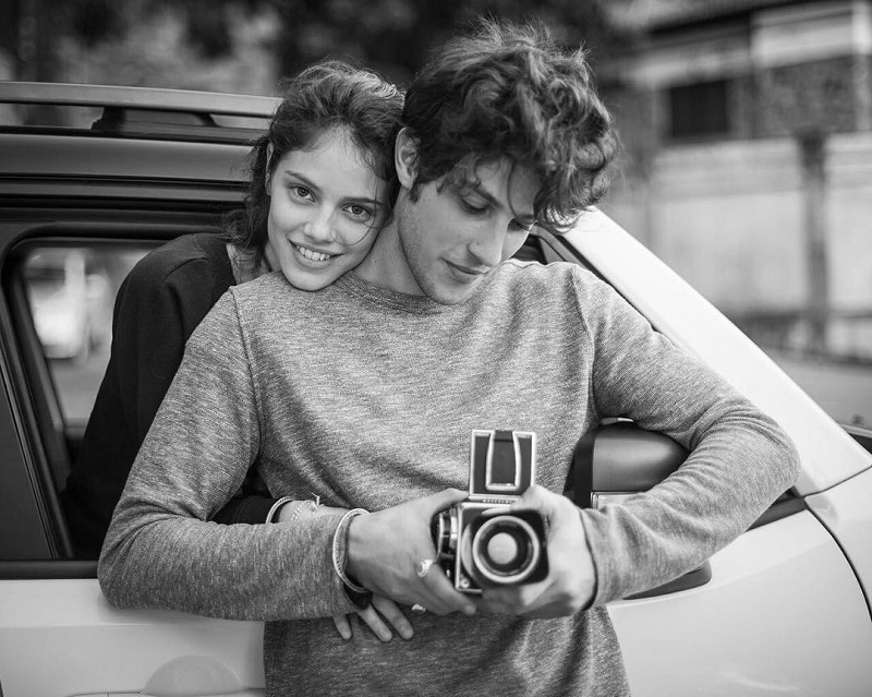 Laura Neiva abraça Chay Suede, que segura uma câmera fotográfica antiga. Em preto e branco.