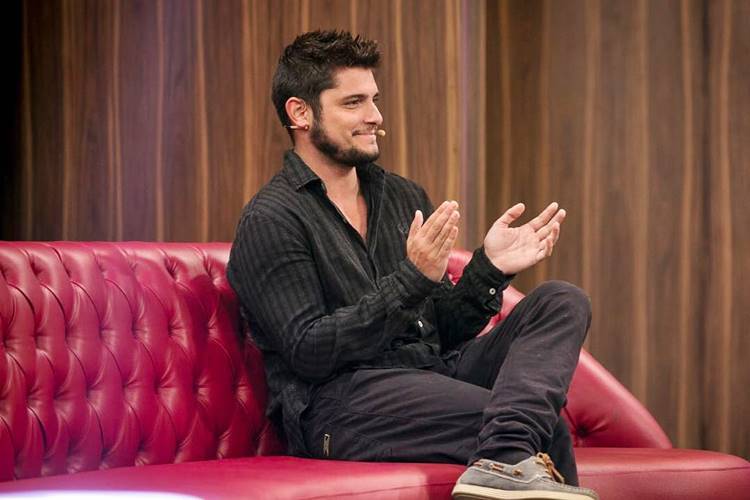 Bruno Gissoni bate palmas sentado no sofá vermelho do estúdio onde é gravado o programa Tamanho Família