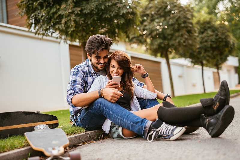 Casal de menina e menino sentados na sarjeta olhando o celular e rindo