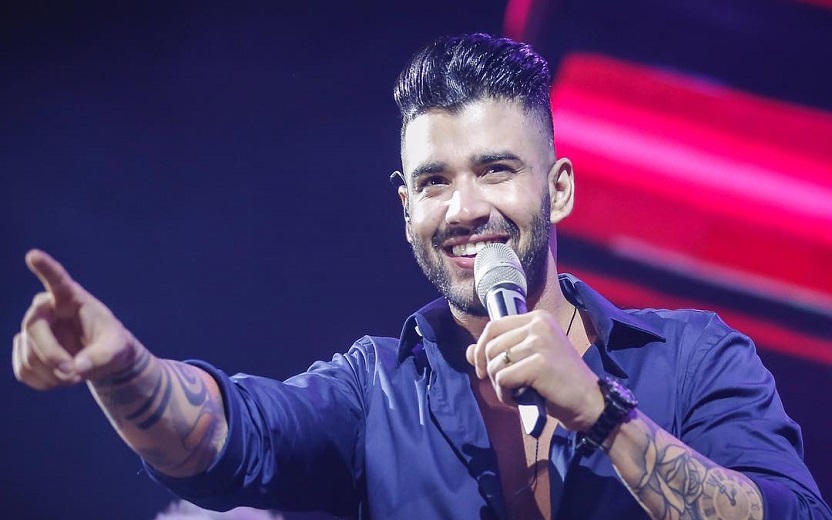 Gusttavo Lima cantando e apontando em show, usando camisa azul