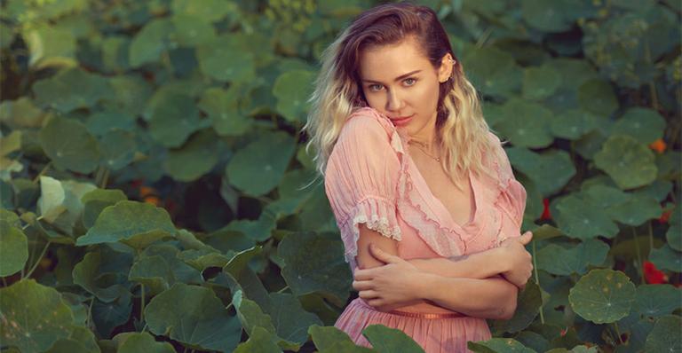 Miley aparece com os cabelos compridos e soltos, usando um vestido rosa claro, em meio a um jardim
