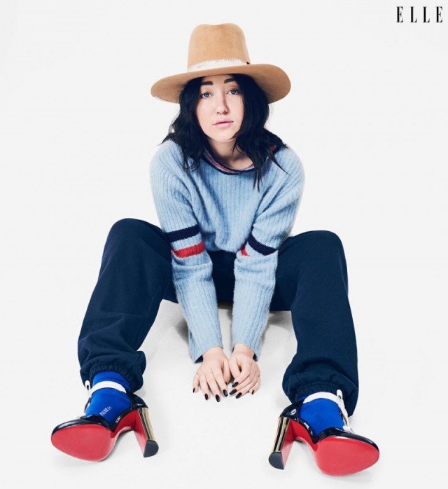 Noah Cyrus sentada no chão usando salto vermelho, meias azuis, moletom e chapéu