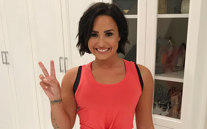 Demi Lovato com camiseta laranja sorrindo e fazendo sinal de "paz e amor"