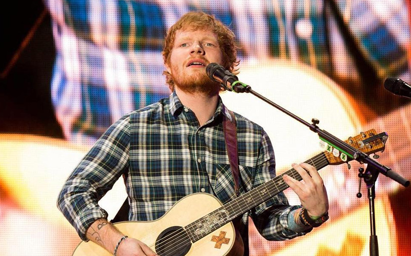 Ed Sheeran toca violão e canta, usando microfone. Ele veste camisa xadrez cinza, Twitter do Ed Sheeran
