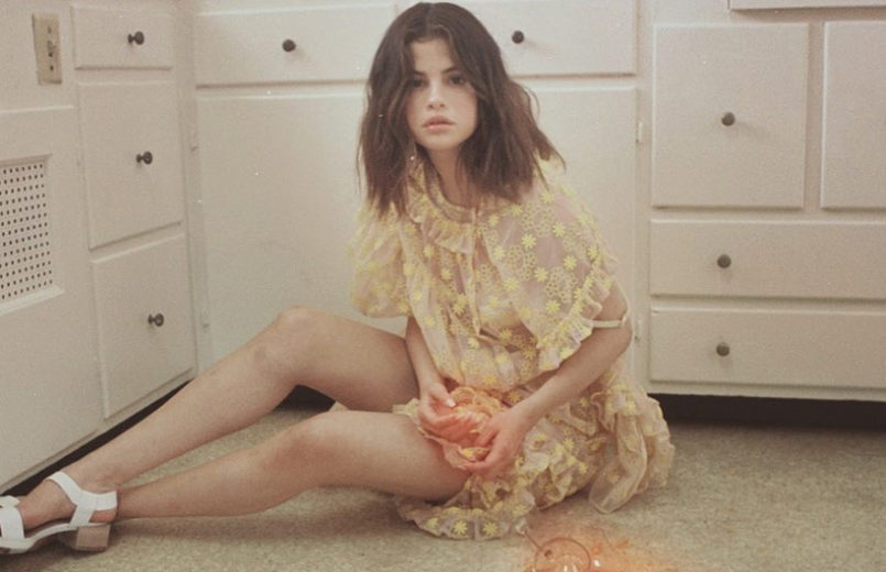 Selena Gomez recolhe cacos de vidro do chão, sentada, usando vestido amarelo transparente