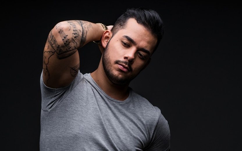 Higor Rocha usa camiseta cinza e exibe tatuagens no braço em fundo preto