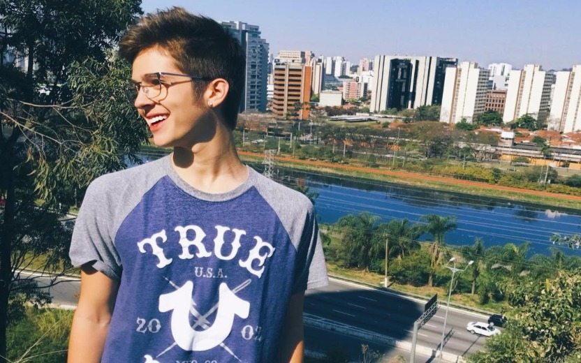João Guilherme sorrindo de perfil em um parque com camiseta azul