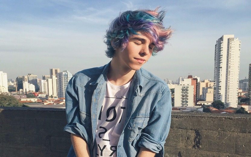 Erick Mafra, menino branco vestindo camisa jeans e de cabelo colorido, de olhos fechados com um fundo de céu e edifícios